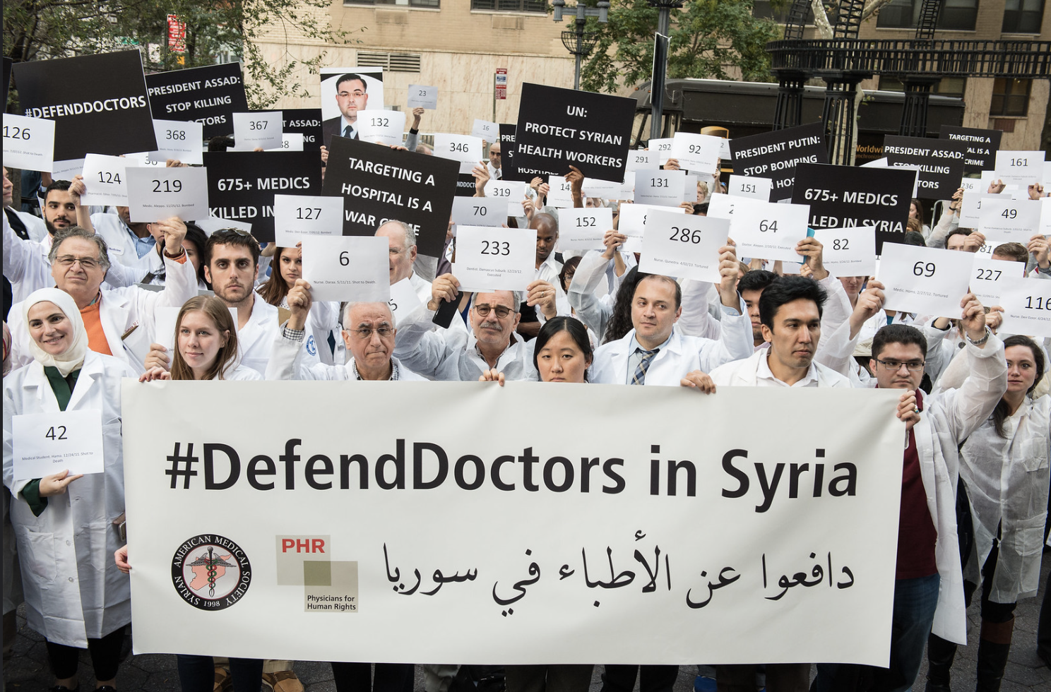 Caption: Medical personnel have been demanding protection for doctors in Syria for years, now this protection is critical to keep the deadly coronavirus from spreading. Here, supporters demand an end to the bombing of hospitals in Syria at a die-in to defend Syrian health professionals, November 4, 2015 (Photo by Michael Hnatov courtesy Physicians for Human Rights) Creative Commons license via Flickr