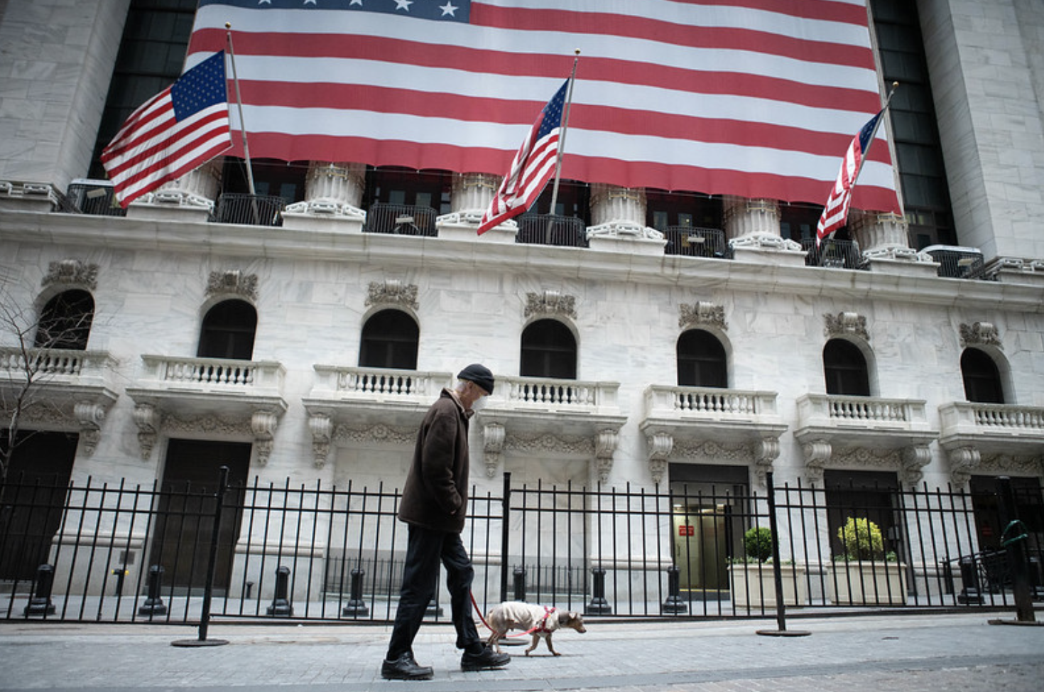 Man in a mask to prevent spread of the coronavirus walks his dog in front of the New York Stock Exchange, where the floor is closed to traders due to the virus. April 12, 2020 (Photo by Anthony Quintano) Creative Commons license via Flickr